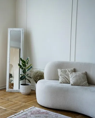 White room with herringbone parquet, a rounded sofa, a tall mirror and sheer curtains